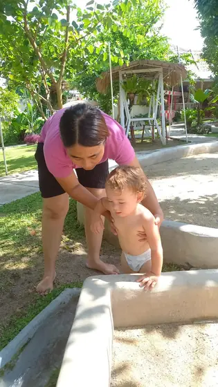 A nanny helping a toddler at a sandpit in a garden